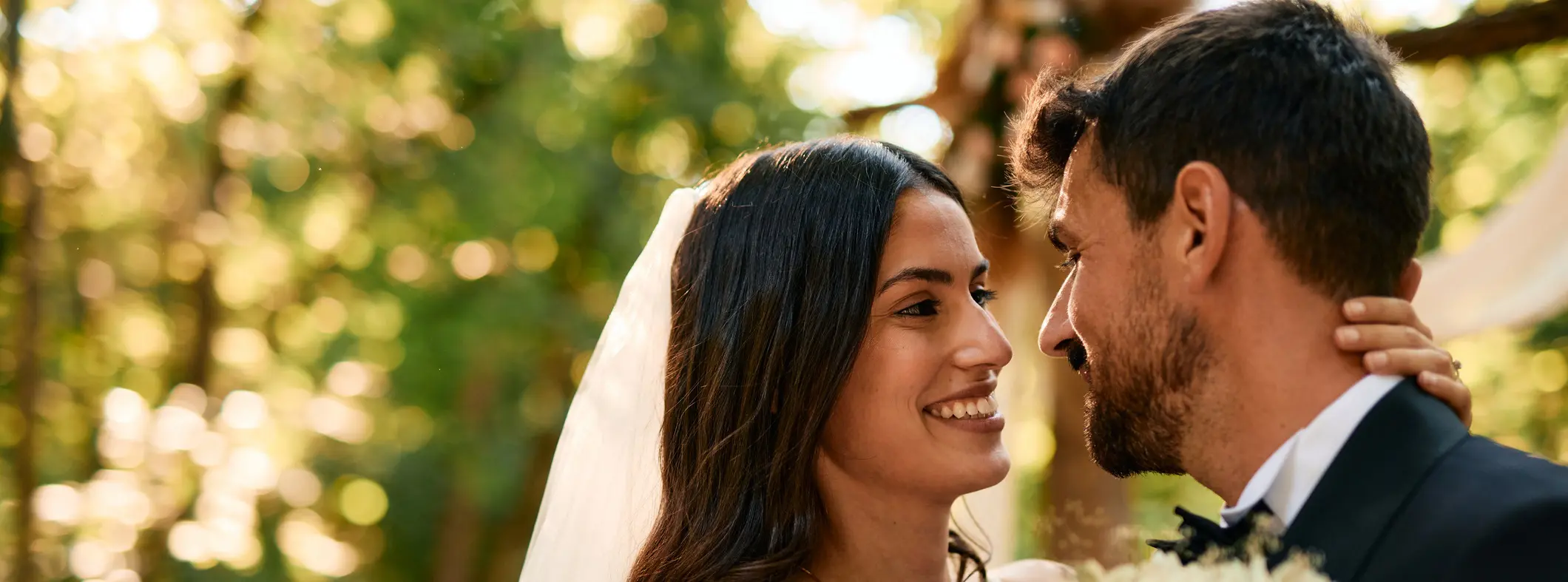 Bride and groom are sharing a tender moment, smiling at each other during their outdoor wedding ceremony, surrounded by the beauty of nature