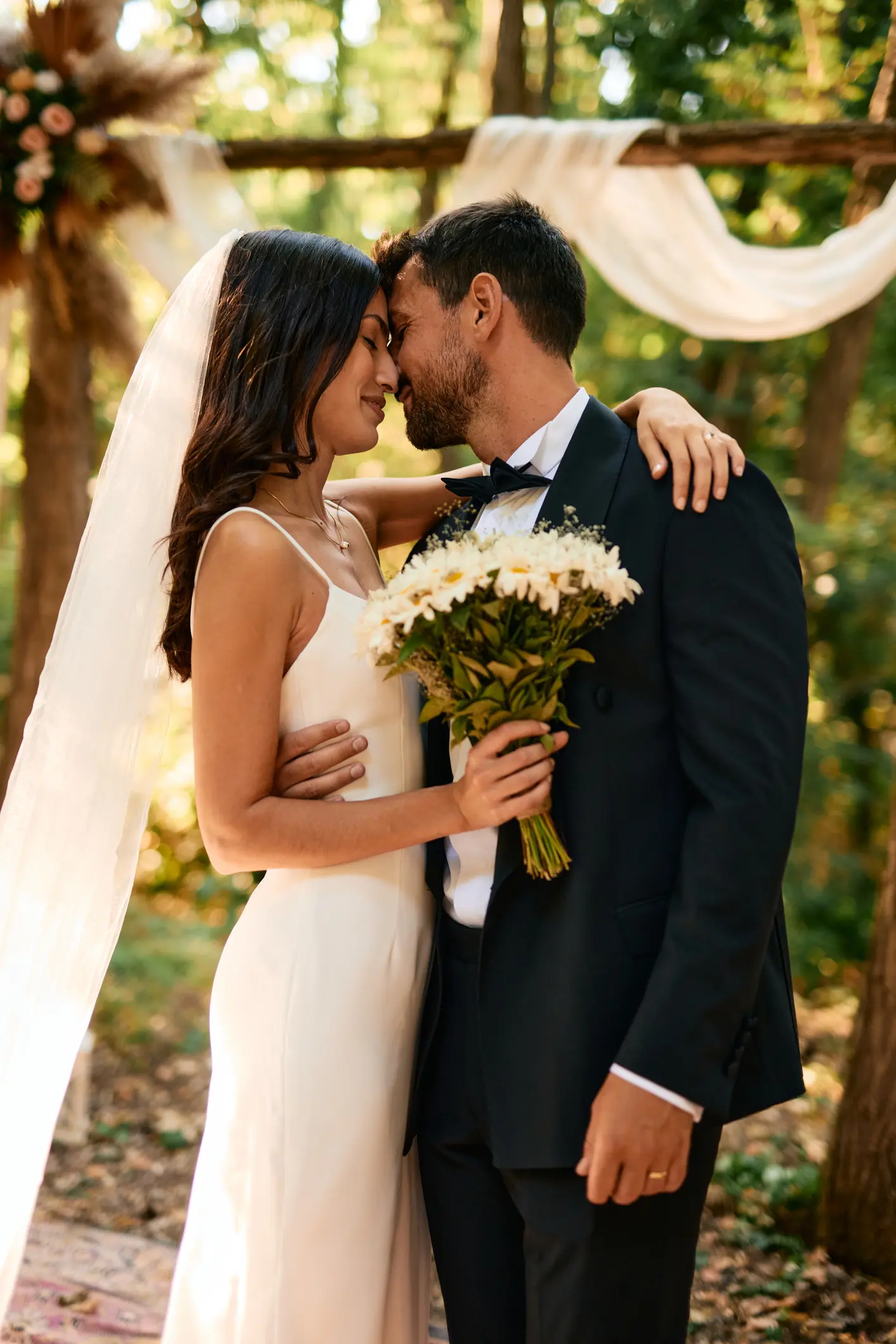 Bride and groom embracing and holding a vibrant bouquet of flowers, celebrating their love during an outdoor wedding ceremony nestled in a serene forest setting